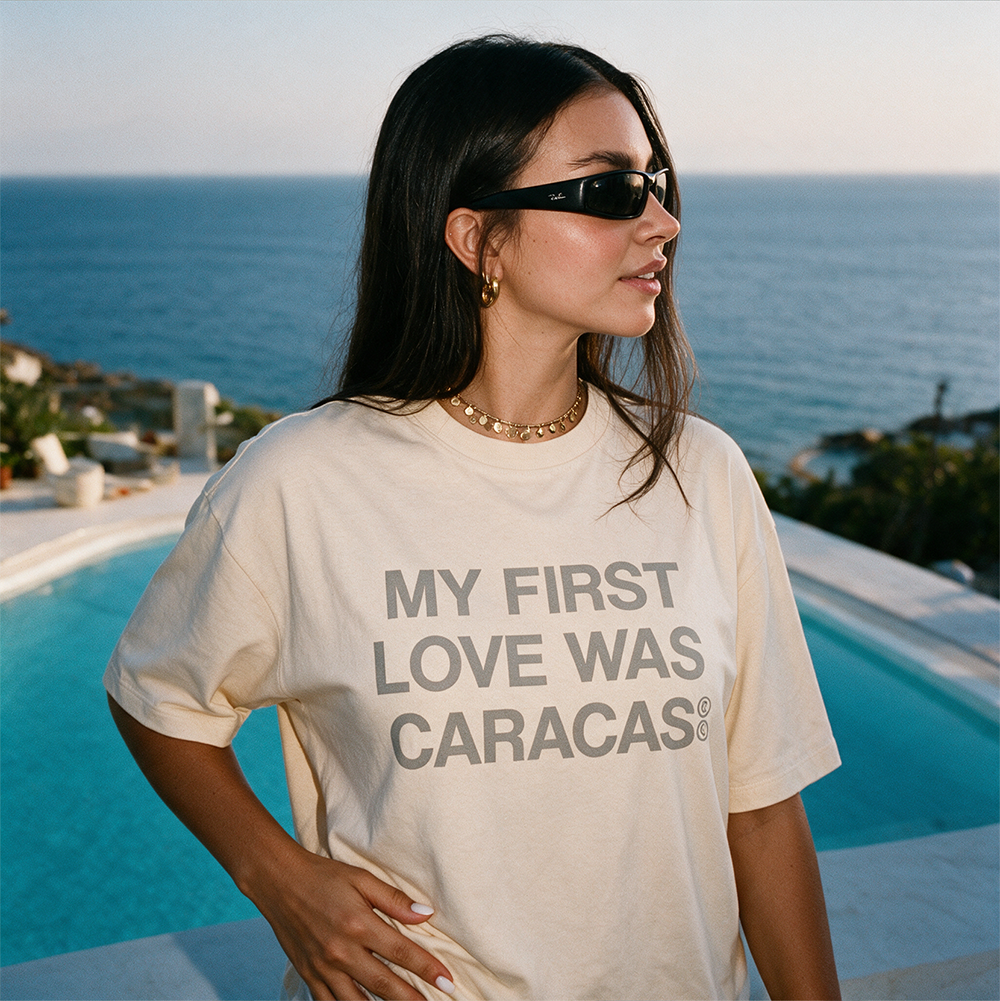 Mujer con gafas de sol llevando la camiseta boxy crema «My First Love Was Caracas» de Caracas Merch junto a una piscina, con el mar al fondo